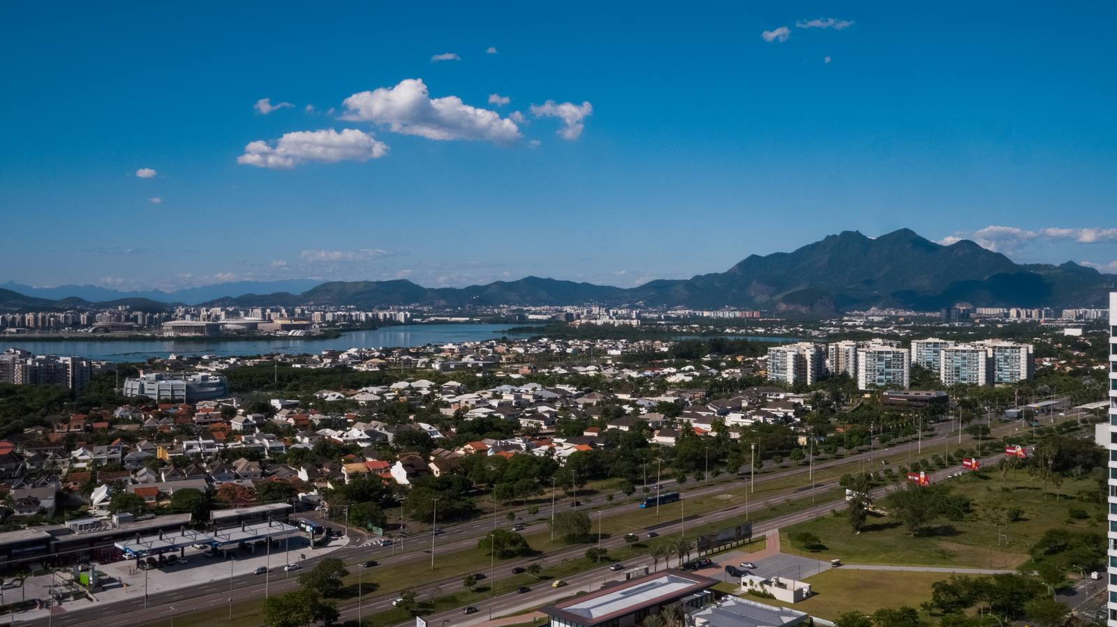 Foto real da Avenida das Américas na região do golfe, com a Lagoa de Jacarepaguá e as montanhas ao fundo